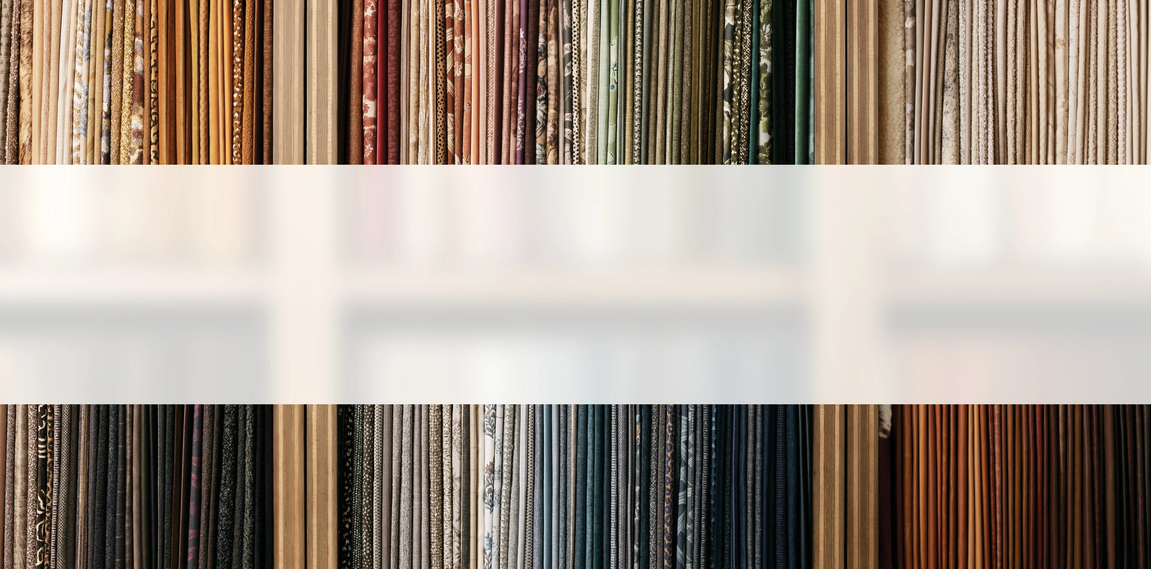 A neatly organized display of vertical hanging fabric samples in a variety of colors and textures, including earthy neutrals, rich jewel tones, and floral patterns. The fabrics are arranged by hue, creating a visually appealing spectrum from light creams and greens to deep blues and browns.